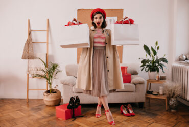 Lady in red beret, beige trench and dress holds shopping bags. Astonished young woman in long skirt and bright heels looks at camera..